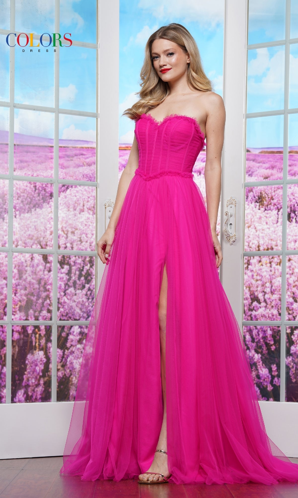 A woman wearing the Colors Dress 3486, a strapless long prom ball gown with a sweetheart neckline and thigh-high slit, stands before glass doors overlooking a lavender field.